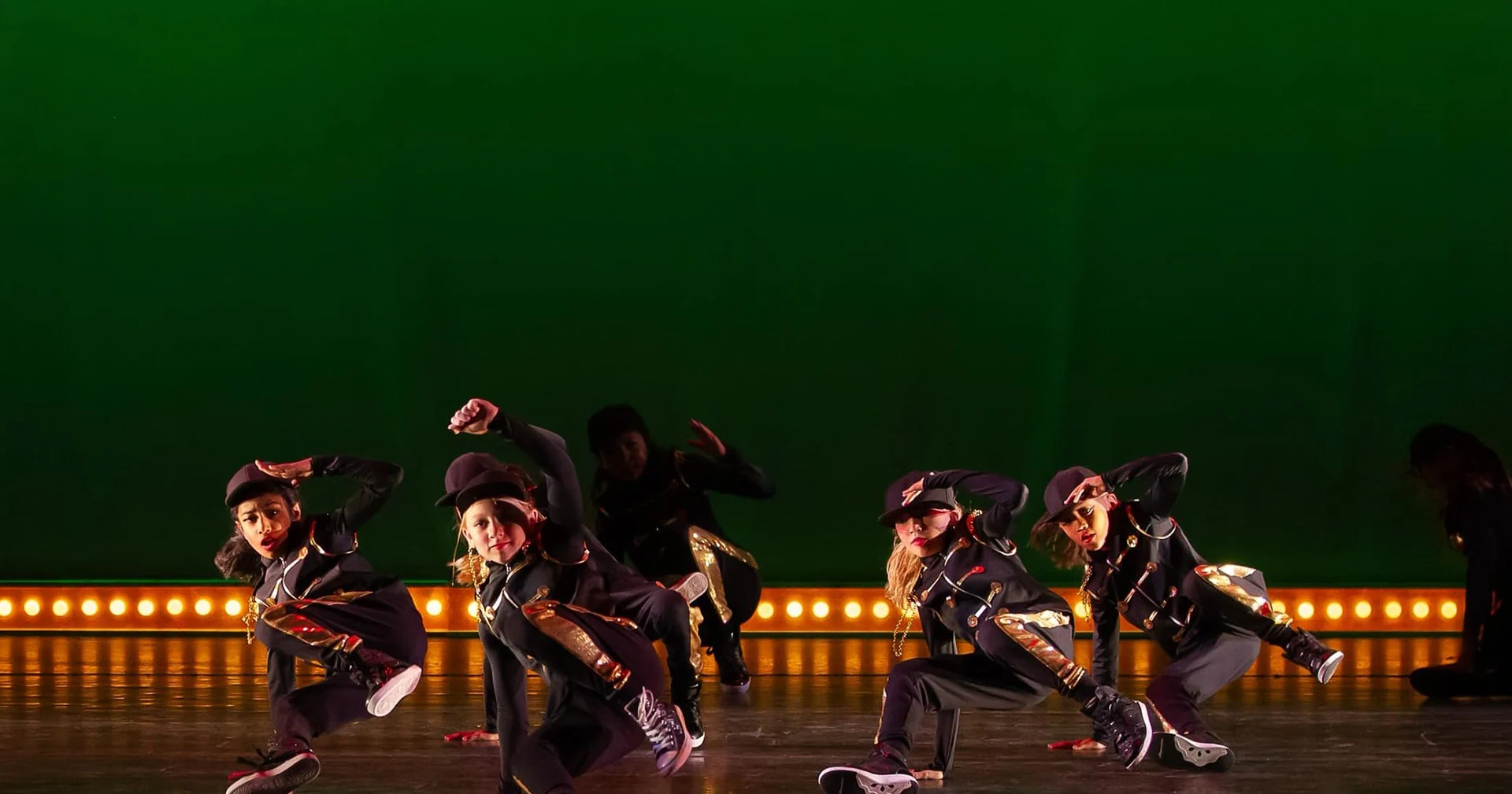 Young hip hop dancers in black and gold military-style jackets crouching in unison on a green-lit stage