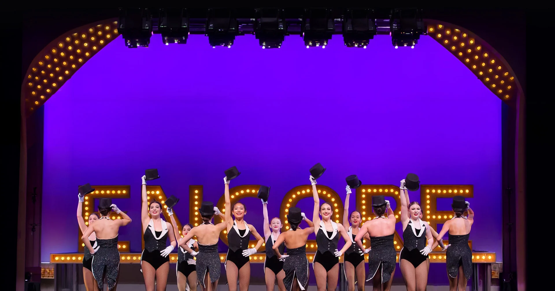 Line of dancers in black tap costumes raising top hats in front of the Encore marquee