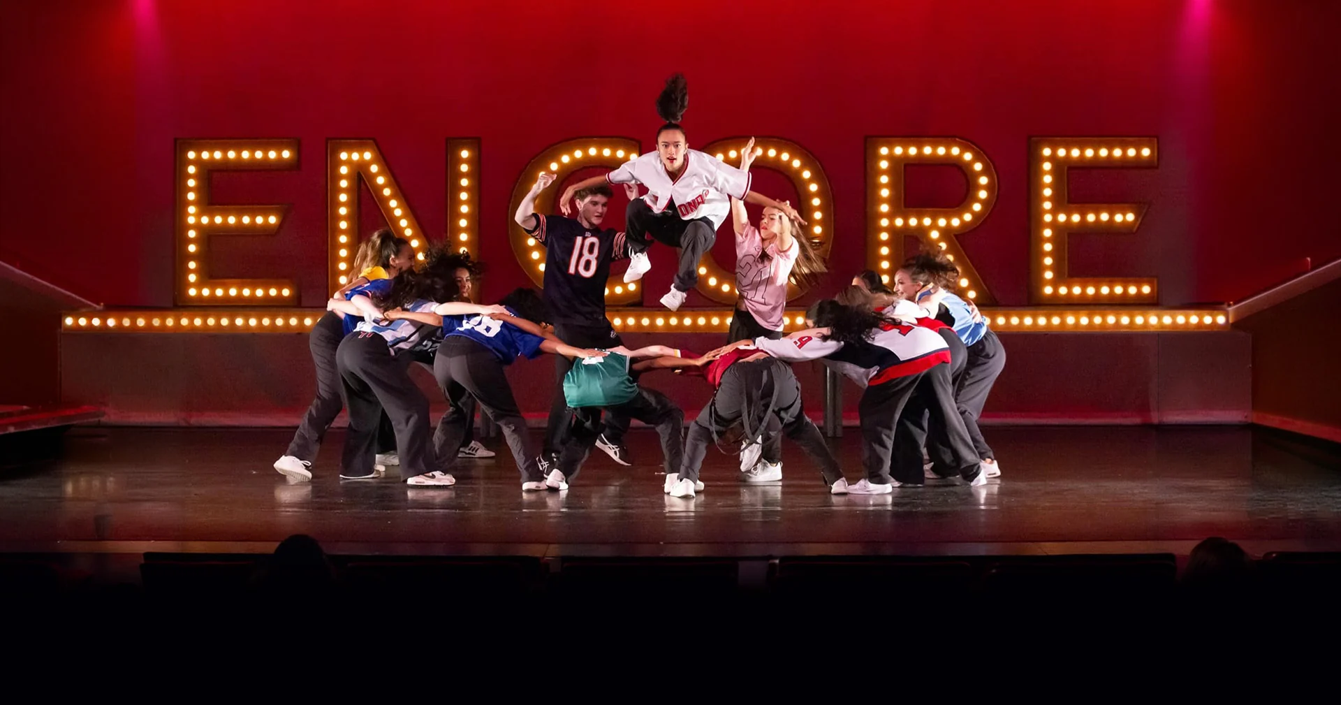 Hip hop crew in sports jerseys mid-routine with one dancer leaping above the group