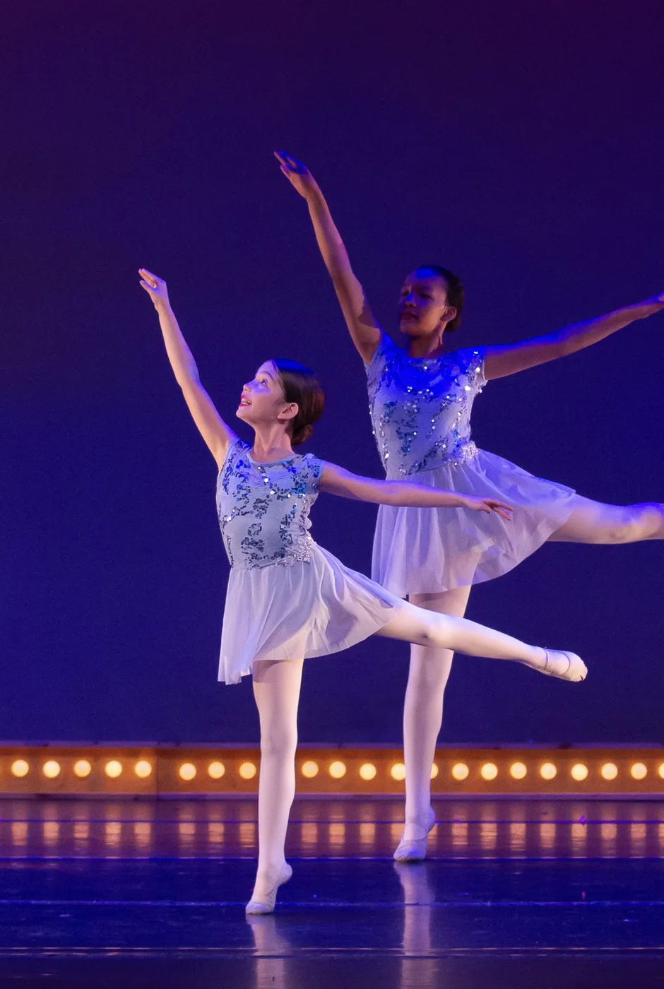 Junior ballet dancers on the blue-lit stage, one holding an arabesque
