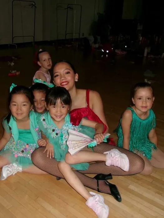 Encore teacher sitting on the studio floor with a group of young dancers in teal costumes