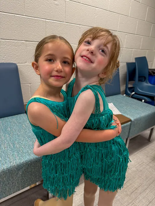 Two young Encore dancers in teal fringe costumes hugging backstage