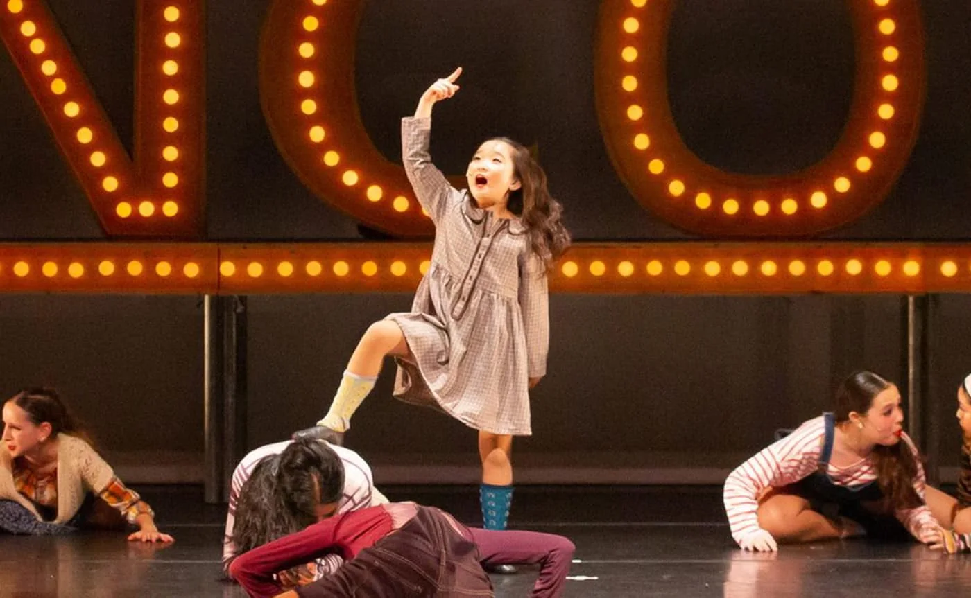 Young Encore dancer pointing skyward mid-performance while dancers pose around her on stage