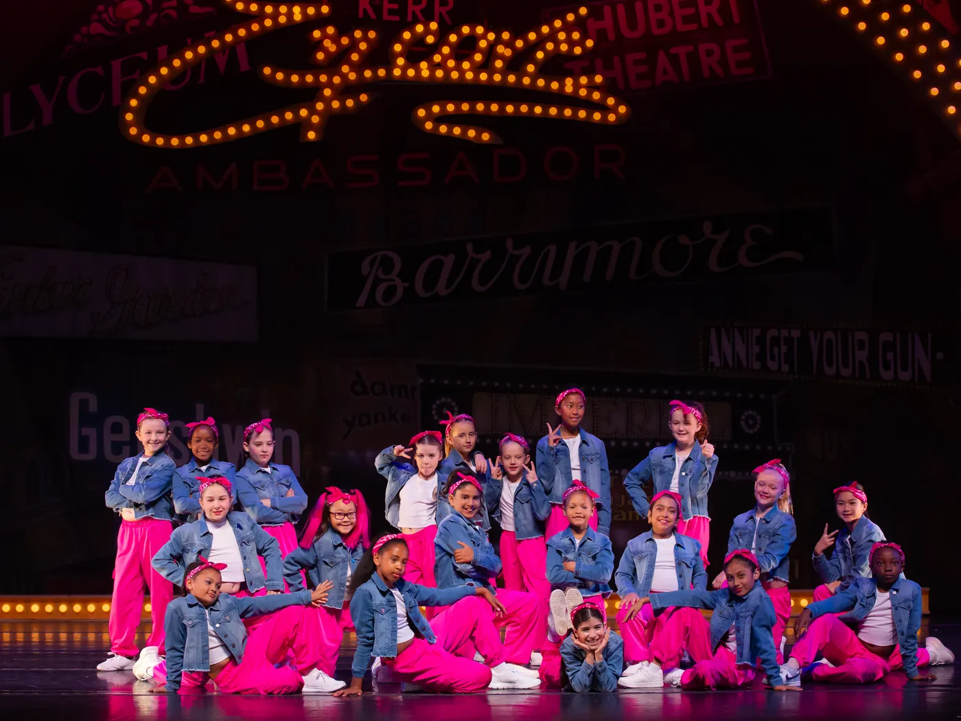 A large group of Encore recreational dancers in denim jackets and pink costumes posed together on stage
