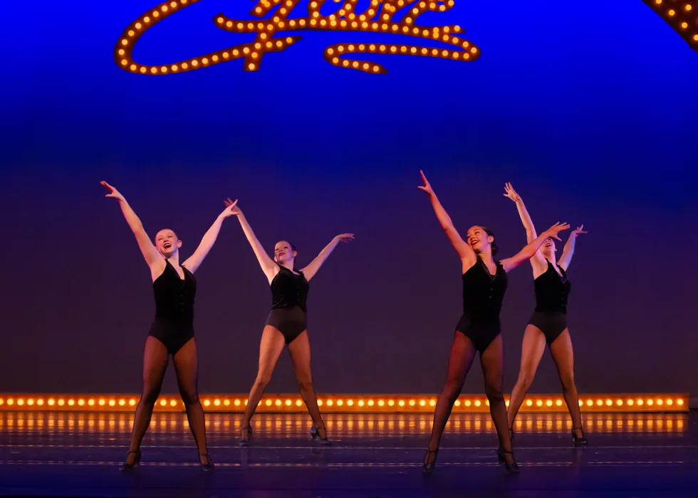 Three Encore dancers reaching upward in unison on a blue-lit stage